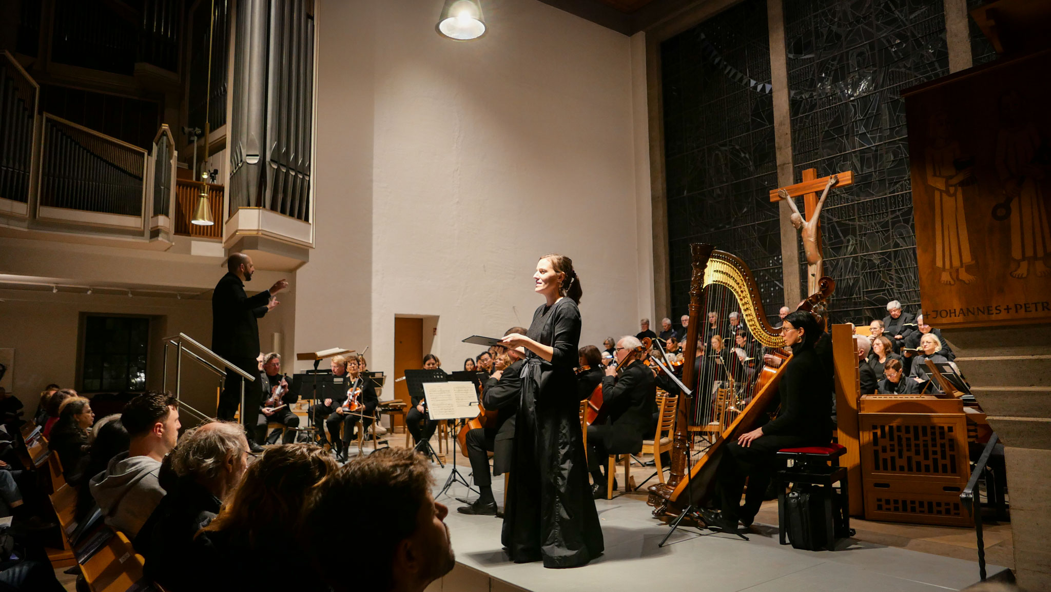 Eine Solisten im schwarzen Kleid singt in einer Kirche. Hinter ihr ein Orchester und sitzend ein gemischter Chor