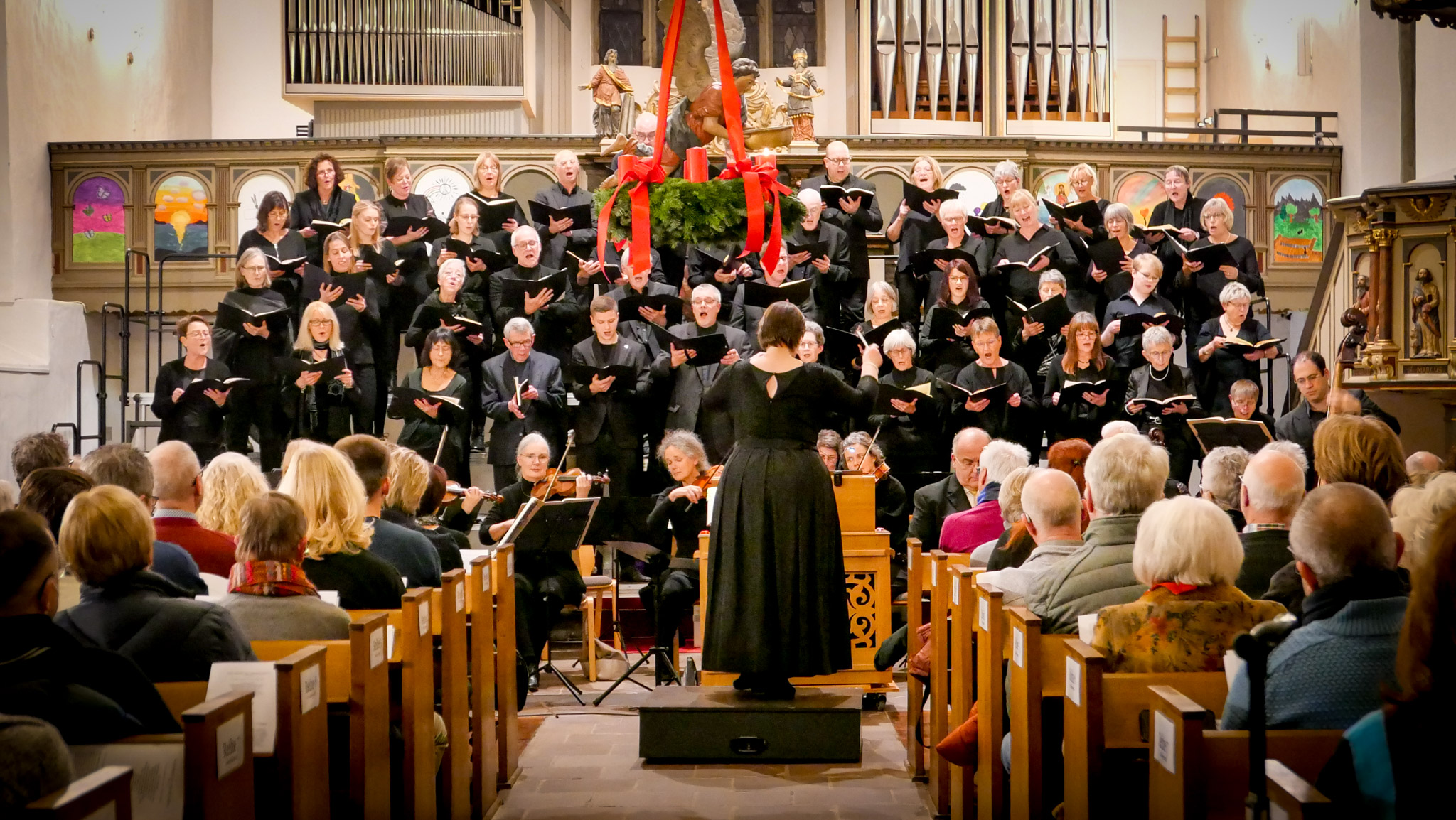 Ein gemischter Chor in Schwarz in einer Kirche, davor eine Dirigentin und ein Orchester, aus dem Mittelgang gesehen